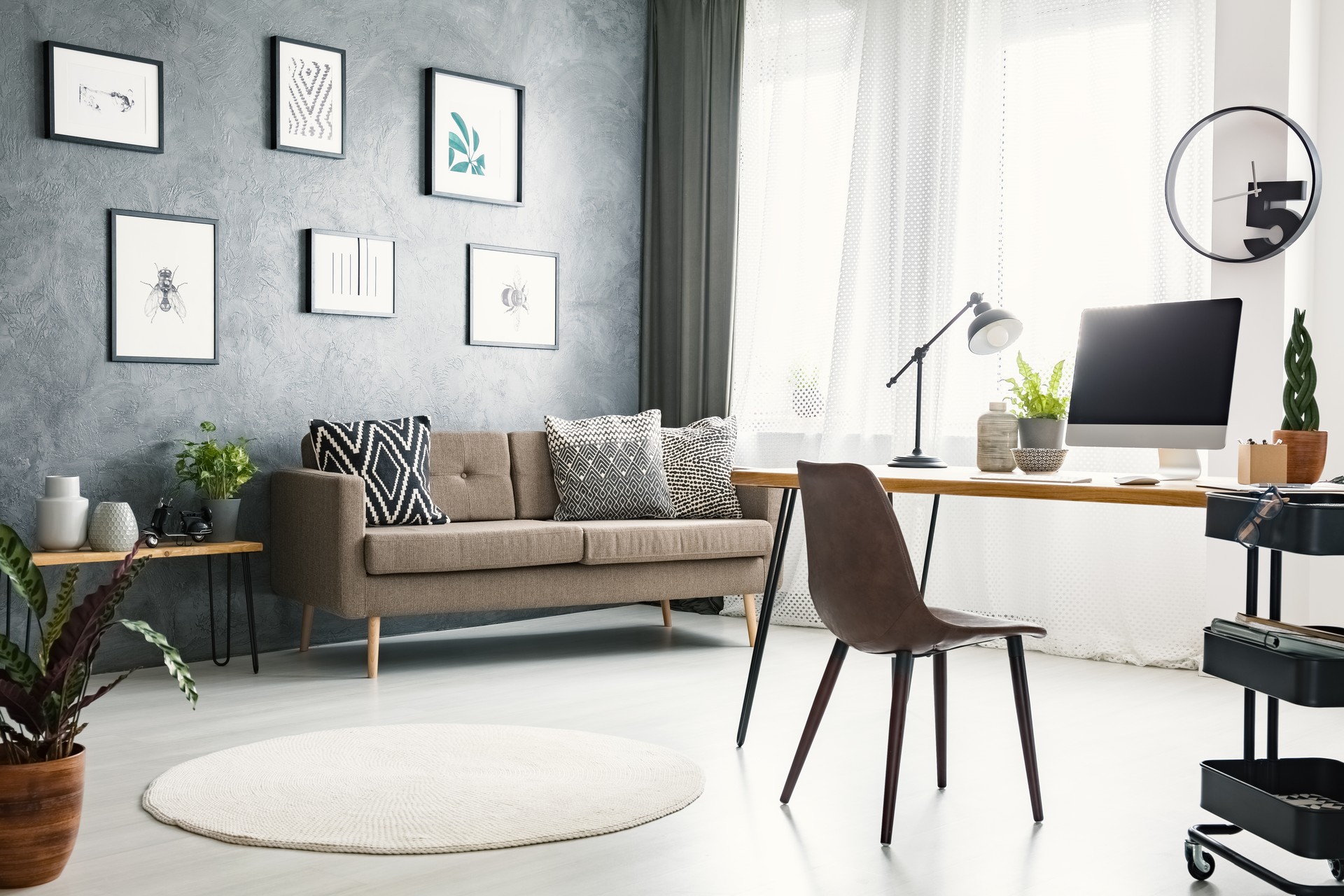 Real photo of a bright home office interior with a sofa, graphics, desk with a computer and lamp and modern clock